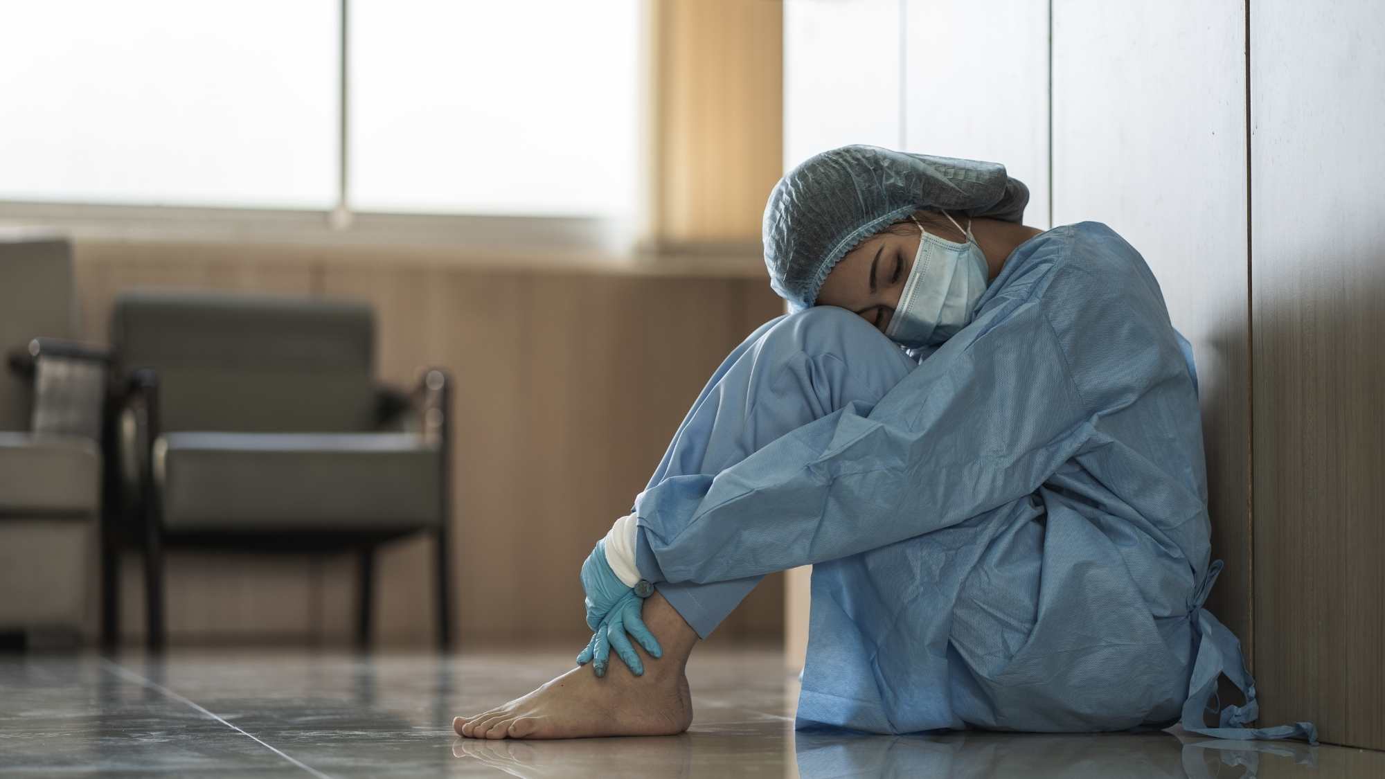 Female health professional in scrubs with mask, barefoot, sitting on floor resting head on knees